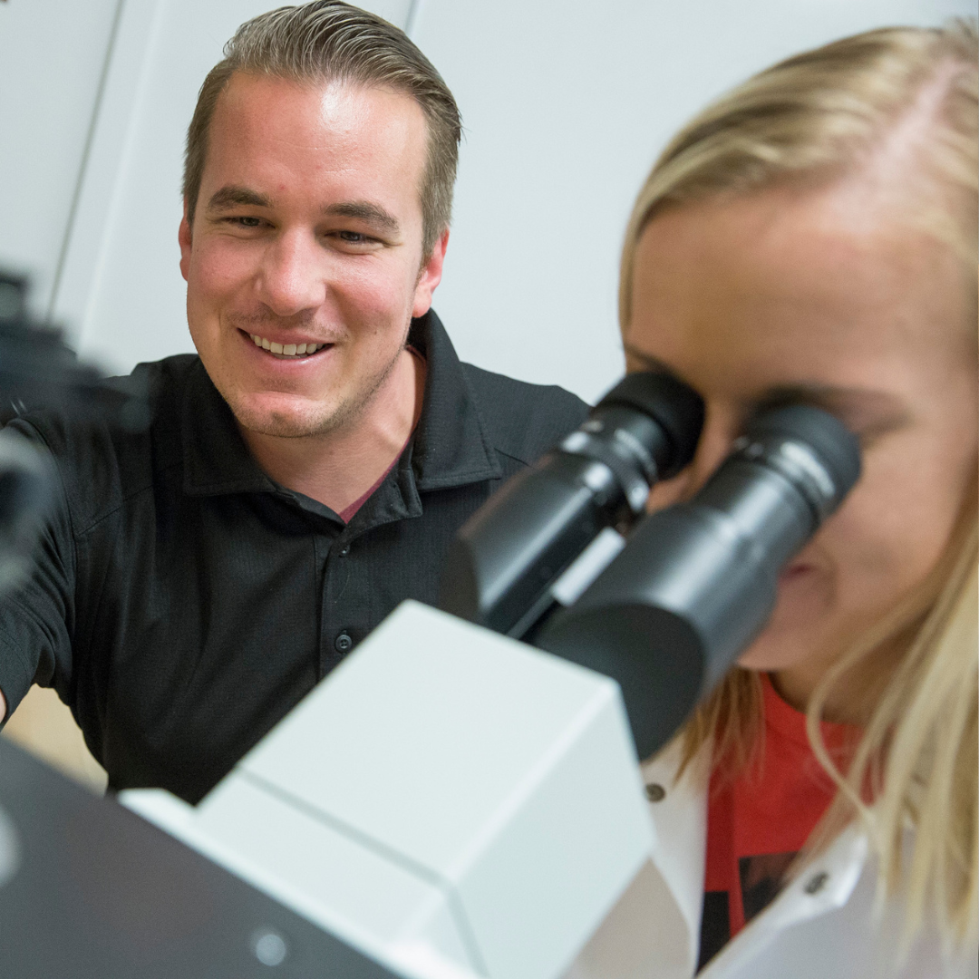 A clinical laboratory scientist looks into a microscope, with another student looking on