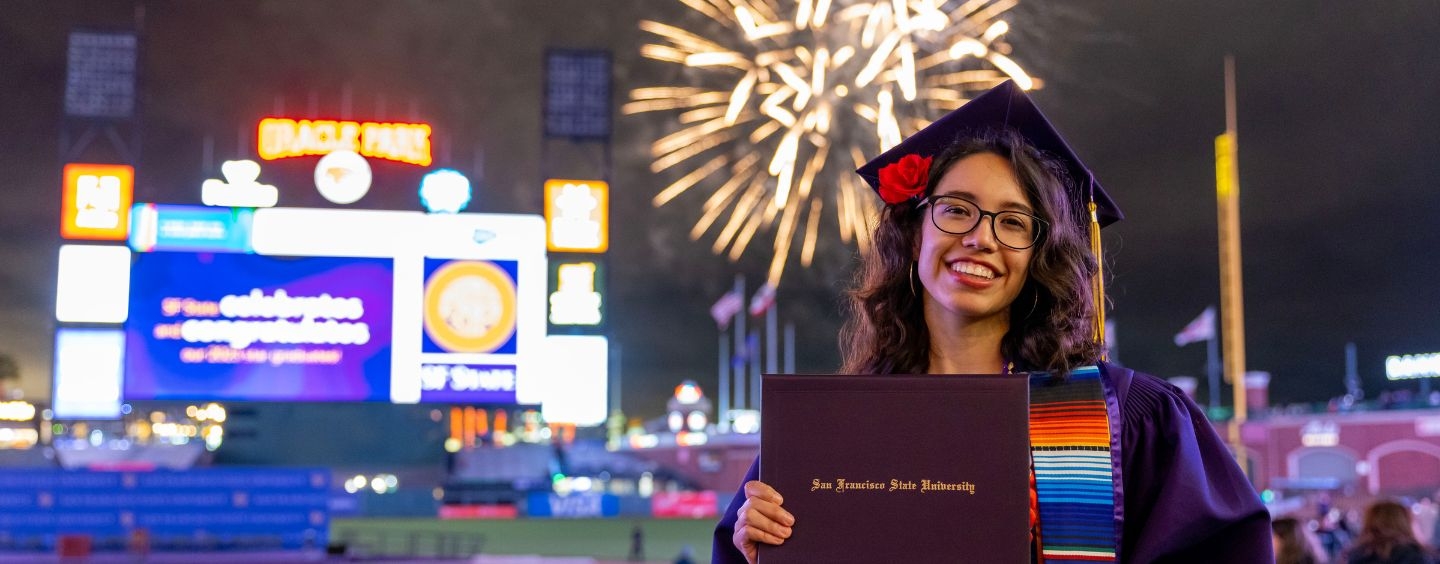 Graduate at Commencement holds up her diploma while fireworks explode at Giants stadium